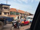 In this picture, you can see a man riding a motorcycle, as well as a red taxi behind him. These types of vehicles are very commonly used all around Ghana
