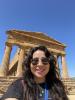 This is a selfie that I took of myself in front of the Temple of Juno in Agrigento, Italy. I have long brown hair, tan skin, and wear sunglasses while smiling in front of a large, ancient Greek temple in the background.